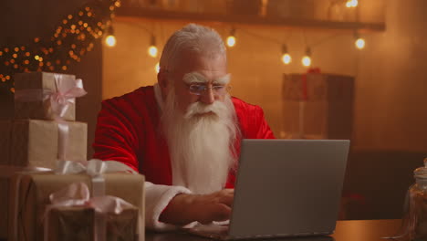 Der-Weihnachtsmann-Hält-Eine-Geschenkbox-In-Der-Hand-Und-Tippt-Auf-Der-Tastatur.-Das-Kind-Spricht-Mit-Dem-Kind-Und-Grüßt-Frohe-Weihnachten-Und-Ein-Frohes-Neues-Jahr-Im-Virtuellen-Online-Chat-Auf-Dem-Laptop.-Am-Weihnachtsabend-Sitzt-Er-Spät-Am-Heimischen-Tisch-Und-Hat-Ein-Geschenk.-Hochwertiges-4K-Filmmaterial