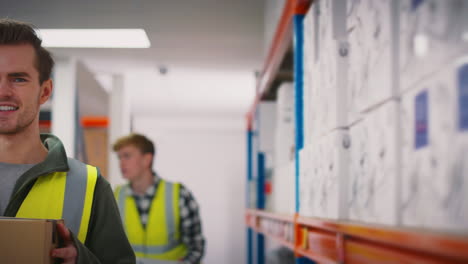 Portrait-Of-Smiling-Male-Worker-Wearing-High-Vis-Safety-Vest-Holding-Box-Inside-Warehouse