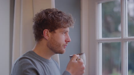 Young-Man-At-Home-Thoughtfully-Looking-Out-Of-Window-And-Drinking-Cup-Of-Coffee