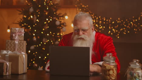 Der-Weihnachtsmann-Hält-Eine-Geschenkbox-In-Der-Hand-Und-Tippt-Auf-Der-Tastatur.-Das-Kind-Spricht-Mit-Dem-Kind-Und-Grüßt-Frohe-Weihnachten-Und-Ein-Frohes-Neues-Jahr-Im-Virtuellen-Online-Chat-Auf-Dem-Laptop.-Am-Weihnachtsabend-Sitzt-Er-Spät-Am-Heimischen-Tisch-Und-Hat-Ein-Geschenk.-Hochwertiges-4K-Filmmaterial