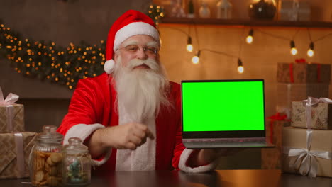 Der-Nette-Weihnachtsmann-Hält-Einen-Laptop-Mit-Grünem-Bildschirm-In-Der-Hand.-Zeigt-Den-Laptop-Bildschirm-An.-Sitzt-In-Einem-Wunderschönen,-Für-Frohe-Weihnachten-Dekorierten-Raum.-Hochwertiges-4K-Filmmaterial
