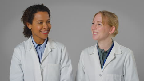 Studio-Portrait-Of-Two-Smiling-Female-Doctors-Or-Lab-Workers-In-White-Coats