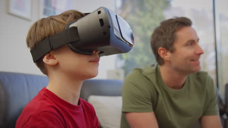 Father-watching-son-wearing-virtual-reality-headset-play-computer-game-sitting-on-sofa-at-home---shot-in-slow-motion