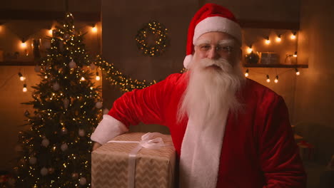 Santa-Claus-stands-in-the-living-room-next-to-a-Christmas-tree-then-takes-a-box-with-a-present-for-an-obedient-child-under-his-armpit-for-a-Happy-Christmas.-Looks-at-the-camera-and-smiles.-High-quality-4k-footage