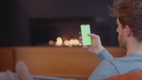 Young-Man-Relaxing-At-Home-Sitting-By-Fire-And-Looking-At-Green-Screen-Mobile-Phone