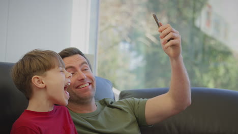 Father-with-son-wearing-pyjamas-sitting-on-sofa-posing-for-selfie-on-mobile-phone---shot-in-slow-motion