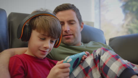 Vater-Mit-Sohn,-Der-Kabellose-Kopfhörer-Trägt-Und-Ein-Computerspiel-Auf-Einem-Handheld-Gerät-Spielt-–-Aufgenommen-In-Zeitlupe