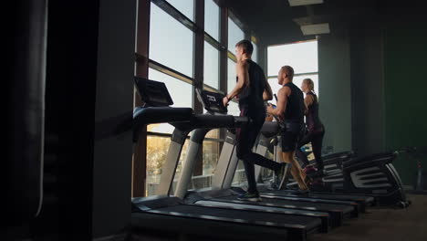 Two-men-running-on-a-treadmill-in-the-background-of-a-large-window-in-the-fitness-room.-Young-men-running-in-the-fitness-room.-Aerobic-workouts-in-the-gym.