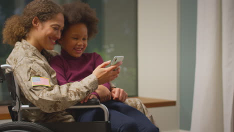 Verletzte-Amerikanische-Soldatin-In-Uniform-Sitzt-Im-Rollstuhl-Und-Schaut-Mit-Ihrer-Tochter-Aufs-Telefon