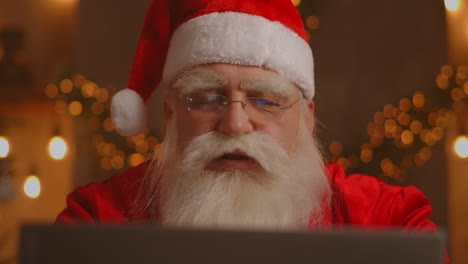 Santa-Claus-holding-gift-box-typing-keyboard-kid-talking-to-child-greeting-on-Merry-Christmas-Happy-New-Year-in-virtual-online-chat-on-laptop-sitting-at-home-table-late-with-present-on-xmas-eve.-High-quality-4k-footage