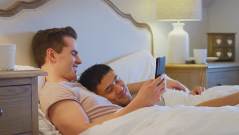 Liebendes-Gleichgeschlechtliches-Männliches-Paar,-Das-Zu-Hause-Im-Bett-Liegt-Und-Sich-Soziale-Medien-Auf-Dem-Handy-Anschaut