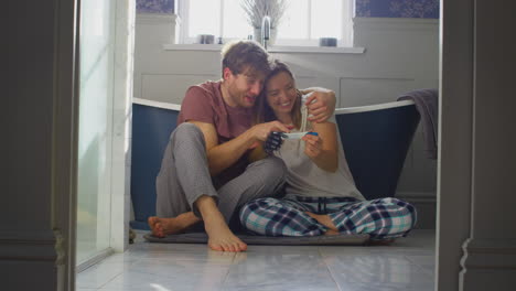 Excited-Couple-With-Woman-With-Prosthetic-Arm-Sitting-On-Bathroom-Floor-With-Positive-Pregnancy-Test
