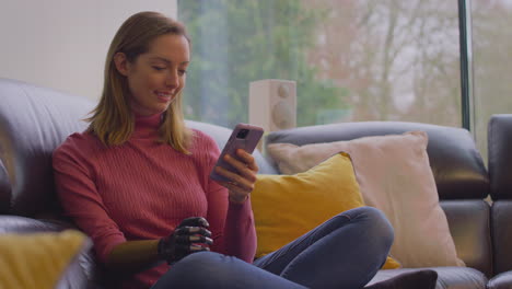 Woman-With-Prosthetic-Arm-On-Sofa-At-Home-Using-Mobile-Phone