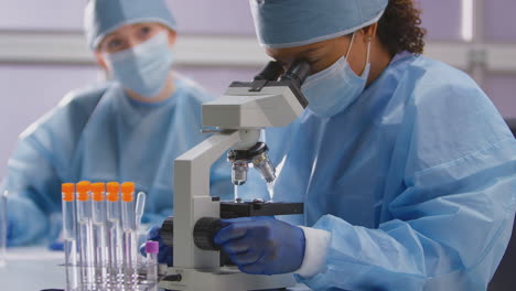 Weibliche-Labormitarbeiter-Tragen-PSA-Und-Analysieren-Proben-Im-Labor-Mit-Mikroskop,-Um-Testergebnisse-Aufzuzeichnen