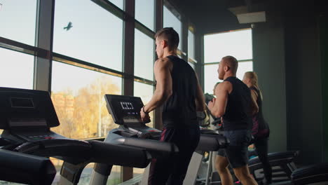 Two-men-running-on-a-treadmill-in-the-background-of-a-large-window-in-the-fitness-room.-Young-men-running-in-the-fitness-room.-Aerobic-workouts-in-the-gym.