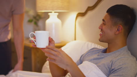 Man-Bringing-Male-Partner-Looking-At-Mobile-Phone-In-Bed-At-Home-Hot-Drink