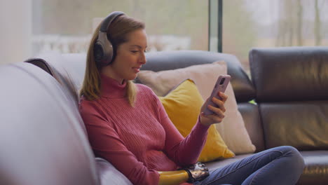Woman-With-Prosthetic-Arm-Wearing-Wireless-Headphones-Watching-Film-On--Mobile-Phone-On-Sofa