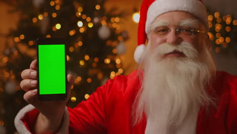Santa-Claus-is-sitting-on-the-sofa-in-the-background-of-a-Christmas-tree-and-garlands-holding-a-mobile-phone-with-a-green-screen-pointing-at-it-with-his-finger.-Santa-holds-a-phone-with-a-chromakey-on-the-screen