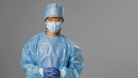 Portrait-Of-Smiling-Female-Surgeon-Wearing-Safety-Glasses-And-Face-Mask-Against-Grey-Background