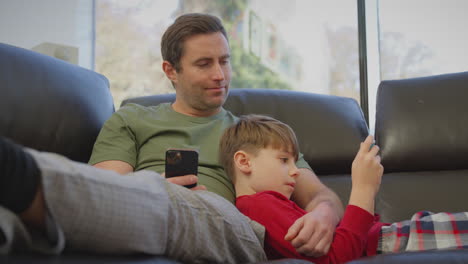 Father-uses-mobile-phone-as-son-plays-computer-game-on-portable-gaming-device-lying-on-sofa-at-home-in-pyjamas---shot-in-slow-motion