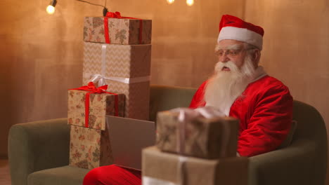 Santa-Claus-video-calling-tak-to-child-on-laptop-hold-present-sit-at-home-table.-Pandemic-and-the-isolation-of-the-Santa-congratulates-children-remotely.-High-quality-4k-footage