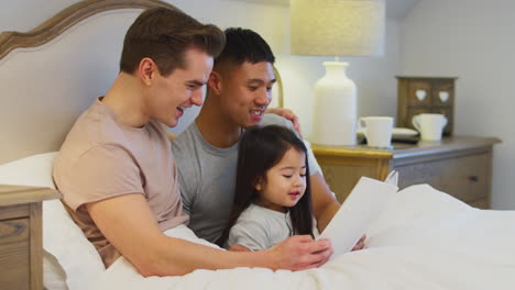 Family-With-Two-Fathers-In-Bed-At-Home-Reading-Story-To-Daughter
