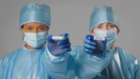 Studio-Shot-Of-Female-Lab-Research-Workers-In-PPE-Holding-Test-Tubes-Labelled-Covid-19-And-Omicron