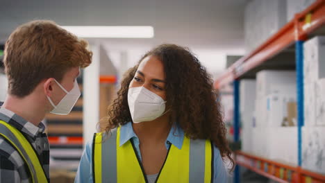 Female-Team-Leader-In-Warehouse-Training-Male-Intern-Standing-By-Shelves-Both-Wearing-Face-Masks