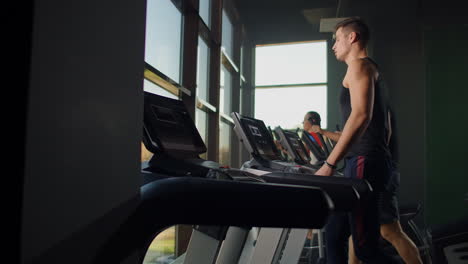 Men-walk-on-treadmills-in-the-fitness-room.-A-group-of-people-walking-on-treadmills-near-a-large-panoramic-window.-Start-of-fitness-classes.-Young-beautiful-women-and-men-in-sportswear-in-cardio-fitness-room