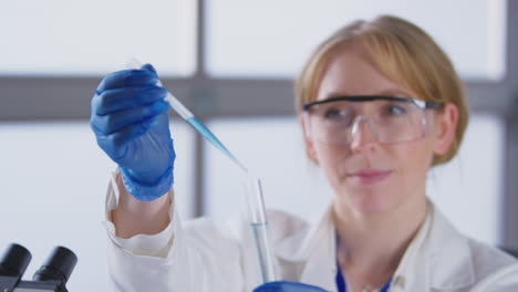 Female-Lab-Worker-Wearing-White-Coat-Adding-Blue-Liquid-From-Pipette-Into-Test-Tube