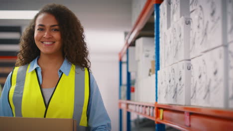 Portrait-Of-Smiling-Female-Worker-Wearing-High-Vis-Safety-Vest-Holding-Box-Inside-Warehouse