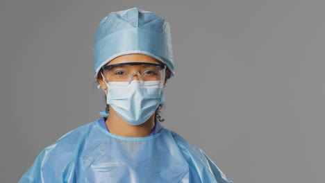 Portrait-Of-Smiling-Female-Surgeon-Wearing-Safety-Glasses-And-Face-Mask-Against-Grey-Background
