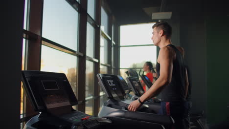Men-walk-on-treadmills-in-the-fitness-room.-A-group-of-people-walking-on-treadmills-near-a-large-panoramic-window.-Start-of-fitness-classes.-Young-beautiful-women-and-men-in-sportswear-in-cardio-fitness-room