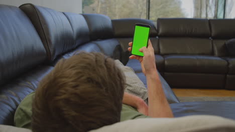 Man-wearing-pyjamas-lying-on-sofa-at-home-looking-at-green-screen-mobile-phone---shot-in-slow-motion