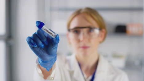 Female-Lab-Research-Worker-Wearing-Safety-Glasses-Holding-Test-Tube-Labelled-Omicron