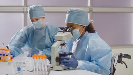 Female-Lab-Worker-In-Wheelchair-Wearing-PPE-Analysing-Samples-In-Laboratory-With-Microscope