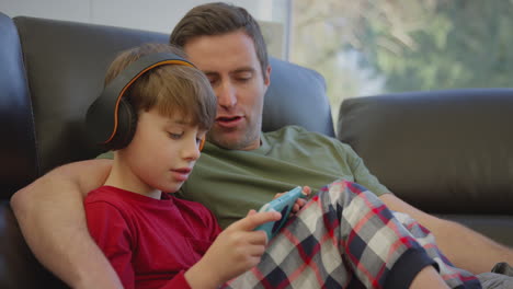 Vater-Mit-Sohn,-Der-Kabellose-Kopfhörer-Trägt-Und-Ein-Computerspiel-Auf-Einem-Handheld-Gerät-Spielt-–-Aufgenommen-In-Zeitlupe