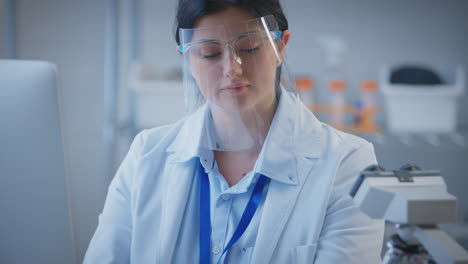 Female-Lab-Worker-Wearing-PPE-Face-Shield-Recording-Test-Results-On-Computer