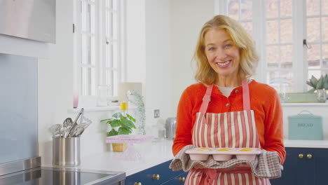 Portrait-Of-Senior-Woman-In-Kitchen-At-Home-Holding-Tray-Of-Freshly-Baked-Cupcakes