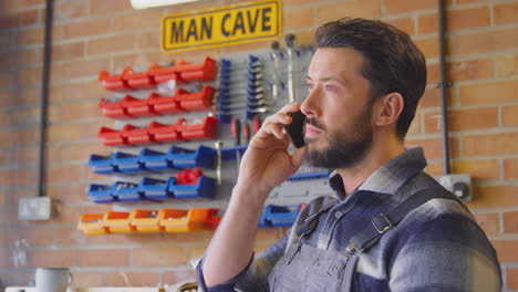 Man-Wearing-Overalls-In-Garage-Workshop-At-Home-Talking-On-Mobile-Phone