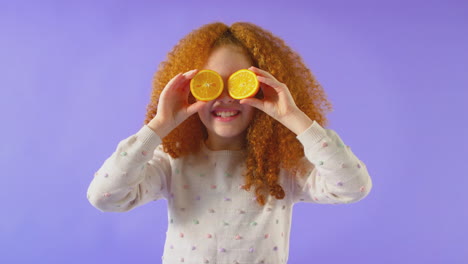 Studioporträt-Eines-Mädchens,-Das-Zwei-Orangefarbene-Hälften-Vor-Violettem-Hintergrund-Vor-Augen-Hält