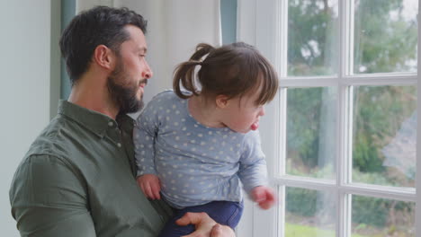 Loving-Father-Holding-Down-Syndrome-Daughter-At-Home-Looking-Out-Of-Window-Together