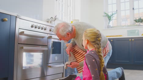 Grandfather-And-Granddaughter-Take-Freshly-Baked-Cupcakes-Out-Of-The-Oven-In-Kitchen-At-Home