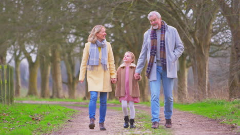 Grandparents-Swinging-Granddaughter-Outside-Walking-Through-Winter-Countryside-Holding-Hands