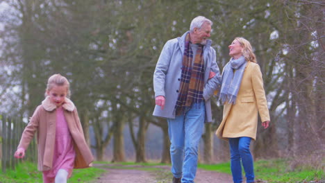 Grandparents-Watching-Granddaughter-Skipping-Ahead-On-Outside-Walking-Through-Winter-Countryside