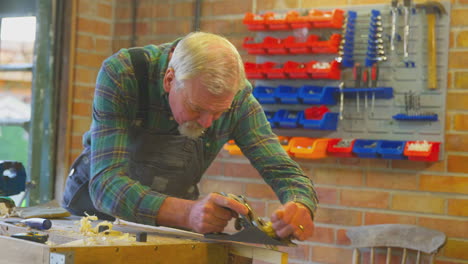 Senior-Male-Carpenter-Wearing-Overalls-In-Garage-Workshop-Planing-Piece-Of-Wood