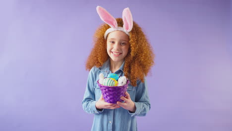Studio-Shot-Of-Girl-Wearing-Rabbit-Ears-And-Holding-Basket-Of-Easter-Eggs-Against-Purple-Background