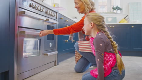 Grandmother-And-Granddaughter-Take-Freshly-Baked-Cupcakes-Out-Of-The-Oven-In-Kitchen-At-Home