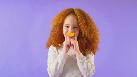 Studio-Portrait-Of-Girl-Holding-Orange-For-Smiling-Mouth-Against-Purple-Background