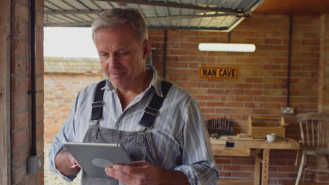 Mature-Male-Wearing-Overalls-Using-Digital-Tablet-In-Garage-Workshop